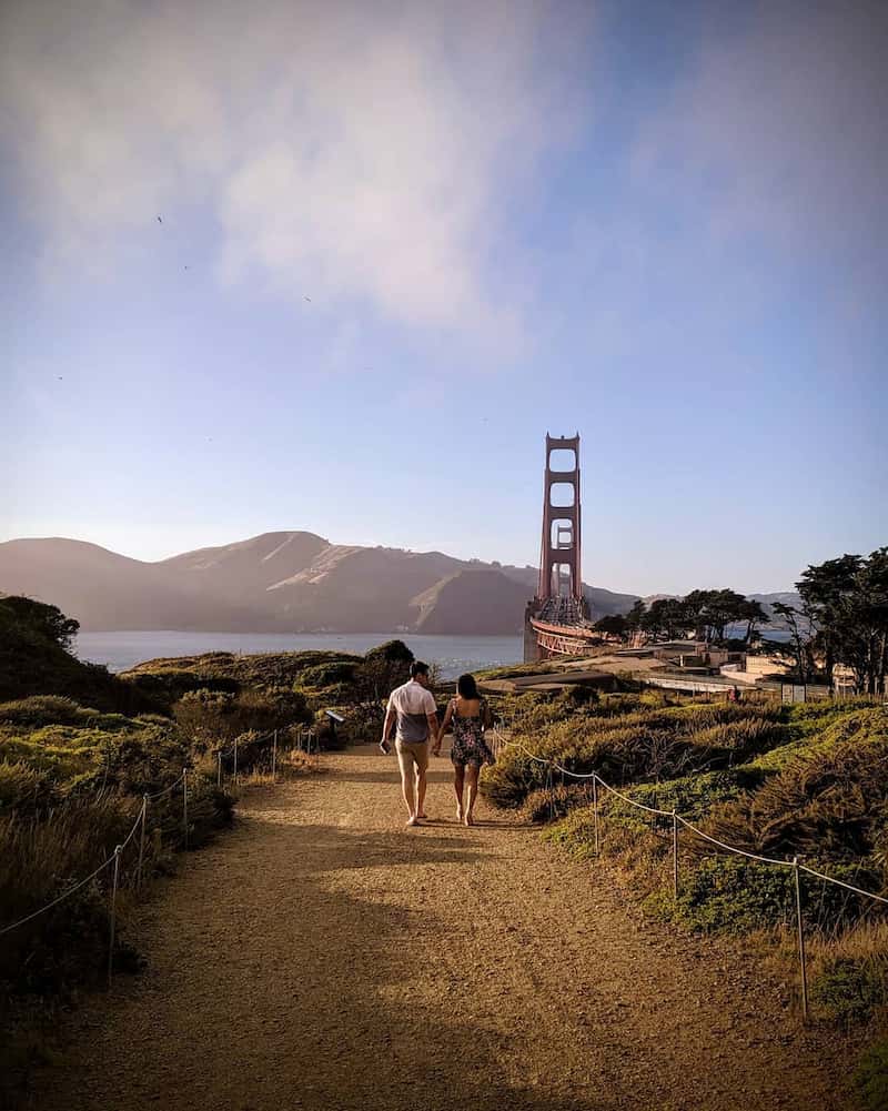 man and woman walking on a dirt path in front of a bridge